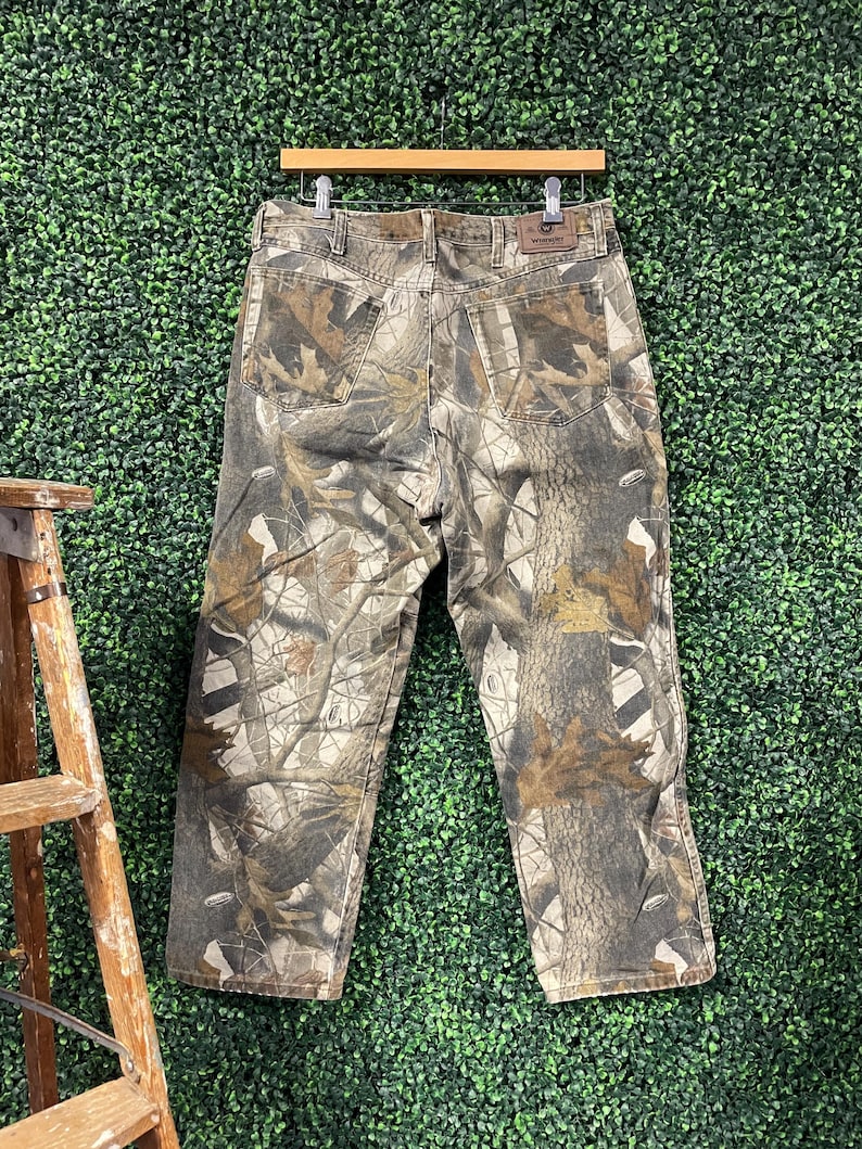 May include: Camouflage-patterned jeans hanging on a wooden hanger. The trousers feature a mix of brown, green, and tan colours, creating a realistic forest design. The jeans have pockets and a leather patch.
