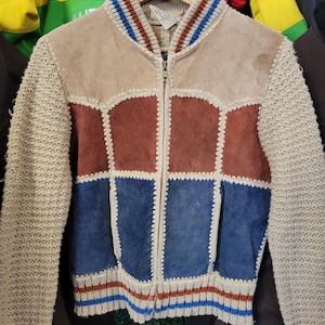 May include: A vintage suede jacket with a patchwork design in tan, brown, and blue. The jacket features knitted sleeves and trim with blue and red stripes. It has a full zipper closure and a classic collar.