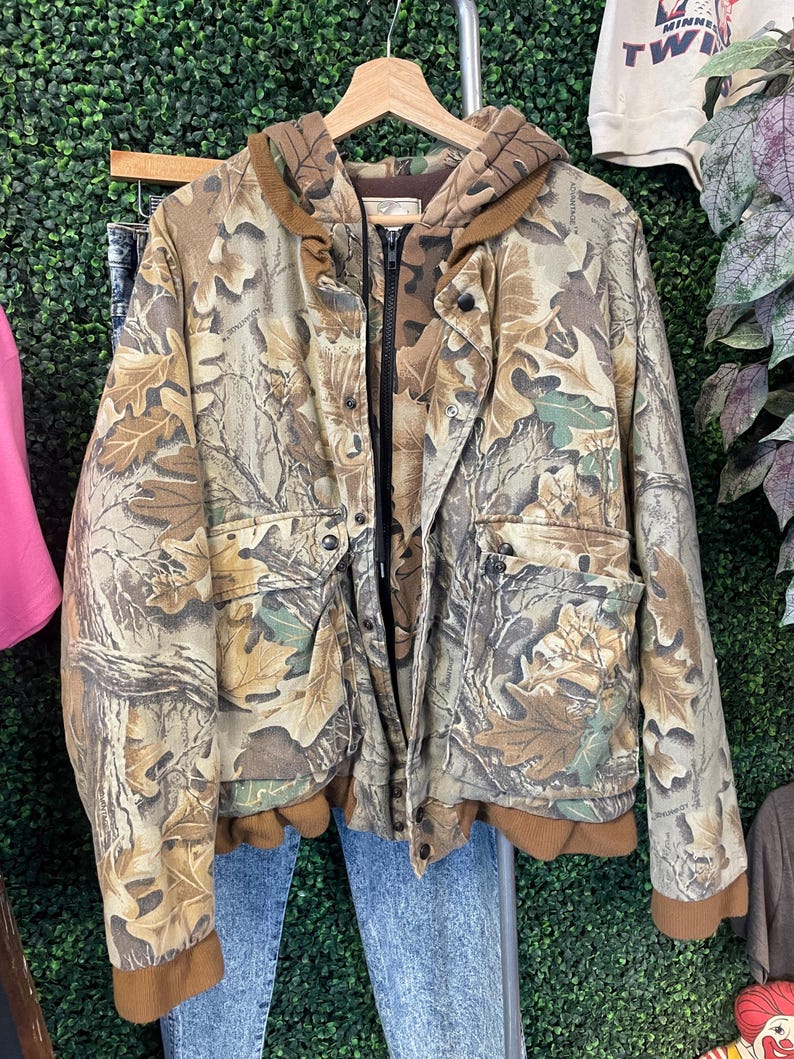 May include: A camouflage jacket with a hood and zipper closure. The jacket features a brown and green leaf pattern. It has two front pockets and brown ribbed cuffs and hem. The jacket is hanging on a wooden hanger against a green background.