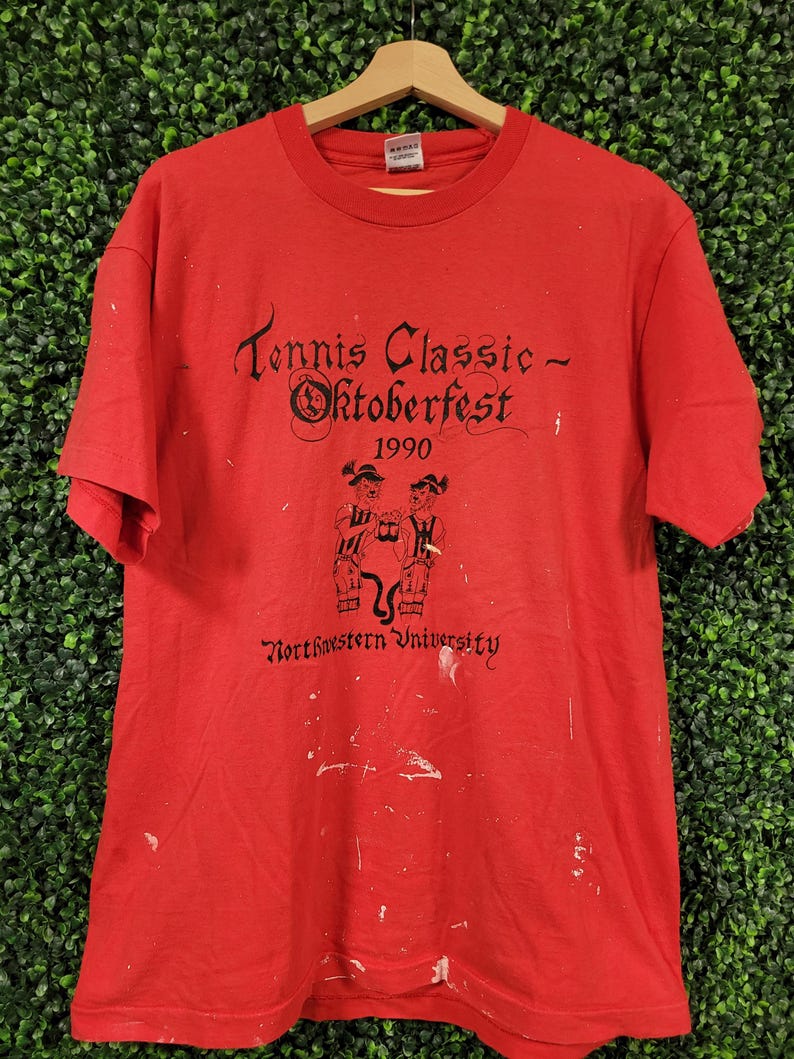May include: Red t-shirt with black text and graphics. The text reads "Tennis Classic - Oktoberfest 1990" with "Northwestern University" below. Features a graphic of two figures. The shirt is hanging on a wooden hanger.