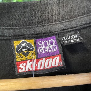 Op de afbeelding: Close-up van een zwart t-shirt met een label met het "Ski-Doo" logo in rood en wit, en "Sno Gear" in paars. Het label bevat ook de maat "TTG/XXL". Het shirt hangt aan een houten hanger.