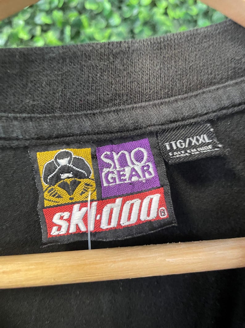 Op de afbeelding: Close-up van een zwart t-shirt met een label met het "Ski-Doo" logo in rood en wit, en "Sno Gear" in paars. Het label bevat ook de maat "TTG/XXL". Het shirt hangt aan een houten hanger.