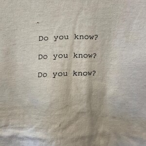 Pu&ograve; includere: T-shirt bianco sporco con la frase "Do you know?" stampata tre volte in nero, centrata verticalmente e orizzontalmente. Il carattere &egrave; semplice, in stile sans-serif.