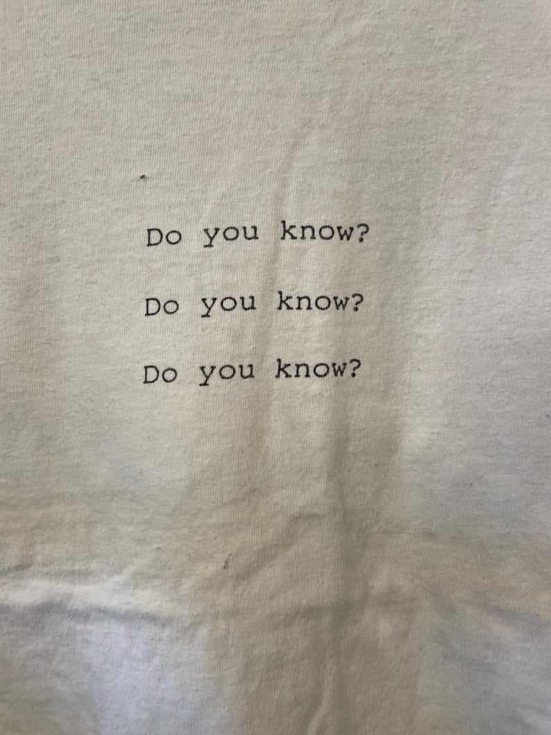 Pu&ograve; includere: T-shirt bianco sporco con la frase "Do you know?" stampata tre volte in nero, centrata verticalmente e orizzontalmente. Il carattere &egrave; semplice, in stile sans-serif.
