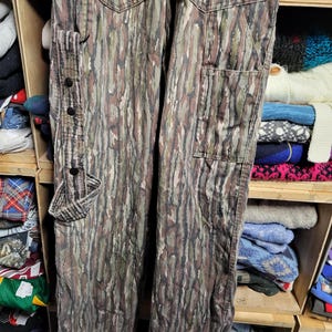 May include: Camouflage-patterned pants in shades of green, brown, and gray. The pants feature multiple pockets, including a cargo pocket on the side and two back pockets. The pants have a buttoned strap detail and are made of a durable fabric.