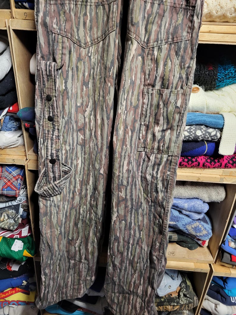 May include: Camouflage-patterned pants in shades of green, brown, and gray. The pants feature multiple pockets, including a cargo pocket on the side and two back pockets. The pants have a buttoned strap detail and are made of a durable fabric.