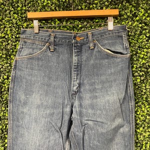 May include: Close-up of blue denim jeans hanging on a wooden hanger. The jeans have a classic five-pocket design with a button and zip closure. The denim has a faded wash, and the jeans are likely vintage.