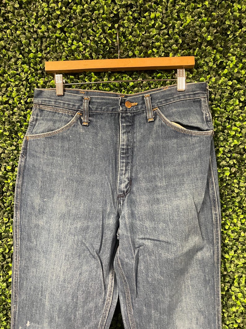May include: Close-up of blue denim jeans hanging on a wooden hanger. The jeans have a classic five-pocket design with a button and zip closure. The denim has a faded wash, and the jeans are likely vintage.