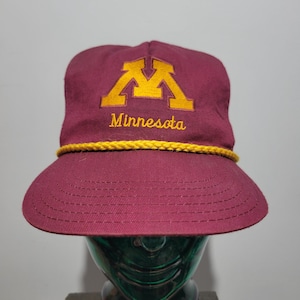 May include: A maroon baseball cap featuring a gold embroidered block M logo and the word Minnesota in gold script. A thick gold braided rope trim sits horizontally across the base of the crown, just above the flat, matching maroon brim.