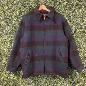 Può includere: Una giacca a maniche lunghe con cerniera e un motivo a quadri scuri nei toni del blu navy, verde bosco e nero. La giacca presenta un colletto standard, una chiusura centrale con zip in metallo e due tasche anteriori inclinate.