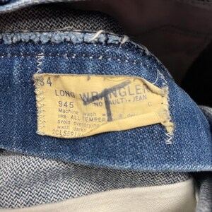 May include: Close-up of blue denim jeans with a yellow label. The label reads "LONG WRANGLER" and includes washing instructions. The jeans are size 34. The fabric is a medium wash denim.