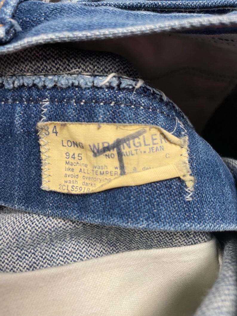 May include: Close-up of blue denim jeans with a yellow label. The label reads "LONG WRANGLER" and includes washing instructions. The jeans are size 34. The fabric is a medium wash denim.