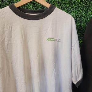 May include: White t-shirt with a dark gray collar and sleeve trim, featuring the text "XBOX 360" in green. The shirt is hanging on a wooden hanger against a green background.