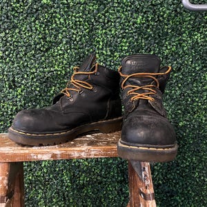 Puede incluir: Par de botas de trabajo de cuero negro con cordones naranjas. Las botas están usadas y están sobre un taburete de madera. Las botas tienen una suela gruesa y la marca es visible en el lateral.