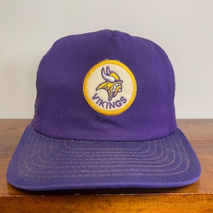 May include: A purple baseball cap featuring a circular patch on the front. The patch has a yellow border and displays a purple and yellow Viking head logo with the text VIKINGS written in purple underneath the graphic.