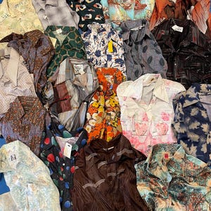 Puede incluir: Una colección de camisas vintage con botones en varios colores y patrones. Las camisas presentan diseños florales, geométricos y abstractos. Algunas camisas tienen etiquetas adjuntas. Las camisas están hechas de diferentes materiales, incluyendo seda y algodón.