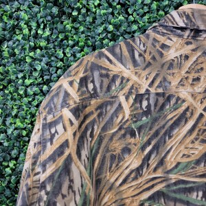 May include: Close-up of a camouflage button-down shirt. The shirt features a pattern of brown, tan, green, and gray, resembling reeds and foliage. The collar and shoulder area are visible, showcasing the fabric's texture and design.