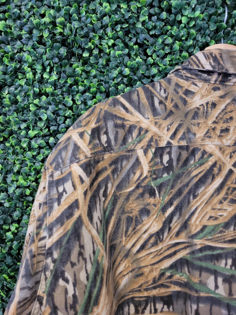 May include: Close-up of a camouflage button-down shirt. The shirt features a pattern of brown, tan, green, and gray, resembling reeds and foliage. The collar and shoulder area are visible, showcasing the fabric's texture and design.