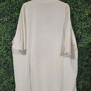 K&ouml;nnte beinhalten: Ein cremefarbenes, &uuml;bergro&szlig;es T-Shirt h&auml;ngt an einem Holzb&uuml;gel vor einem gr&uuml;nen Hintergrund. Das Shirt hat kurze &Auml;rmel und einen Rundhalsausschnitt. Der Stoff scheint leicht zerknittert zu sein. Es gibt einige schwache Flecken auf dem unteren Teil des Shirts.