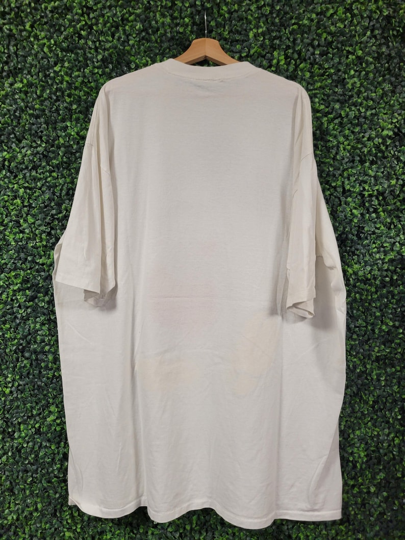 K&ouml;nnte beinhalten: Ein cremefarbenes, &uuml;bergro&szlig;es T-Shirt h&auml;ngt an einem Holzb&uuml;gel vor einem gr&uuml;nen Hintergrund. Das Shirt hat kurze &Auml;rmel und einen Rundhalsausschnitt. Der Stoff scheint leicht zerknittert zu sein. Es gibt einige schwache Flecken auf dem unteren Teil des Shirts.