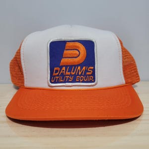 May include: Orange and white trucker hat with a square patch featuring the text "DALUM'S UTILITY EQUIP." The patch has a blue background with an orange "D" logo. The hat has an orange brim and mesh sides.