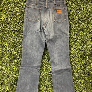 May include: Blue denim Wrangler jeans, viewed from the back, hanging on a wooden hanger. The jeans have two back pockets and a brown leather patch with the Wrangler logo. The jeans have a classic bootcut leg opening.