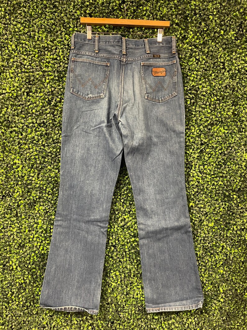 May include: Blue denim Wrangler jeans, viewed from the back, hanging on a wooden hanger. The jeans have two back pockets and a brown leather patch with the Wrangler logo. The jeans have a classic bootcut leg opening.
