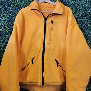 May include: A bright yellow fleece jacket featuring a full-length black front zipper, a high collar, and long sleeves. The garment includes two slanted zippered hand pockets and a single vertical zippered chest pocket on the left side.