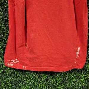 May include: A red t-shirt with white paint marks near the bottom hem. The shirt has a list of text printed on the upper portion. The text includes names of businesses and locations.