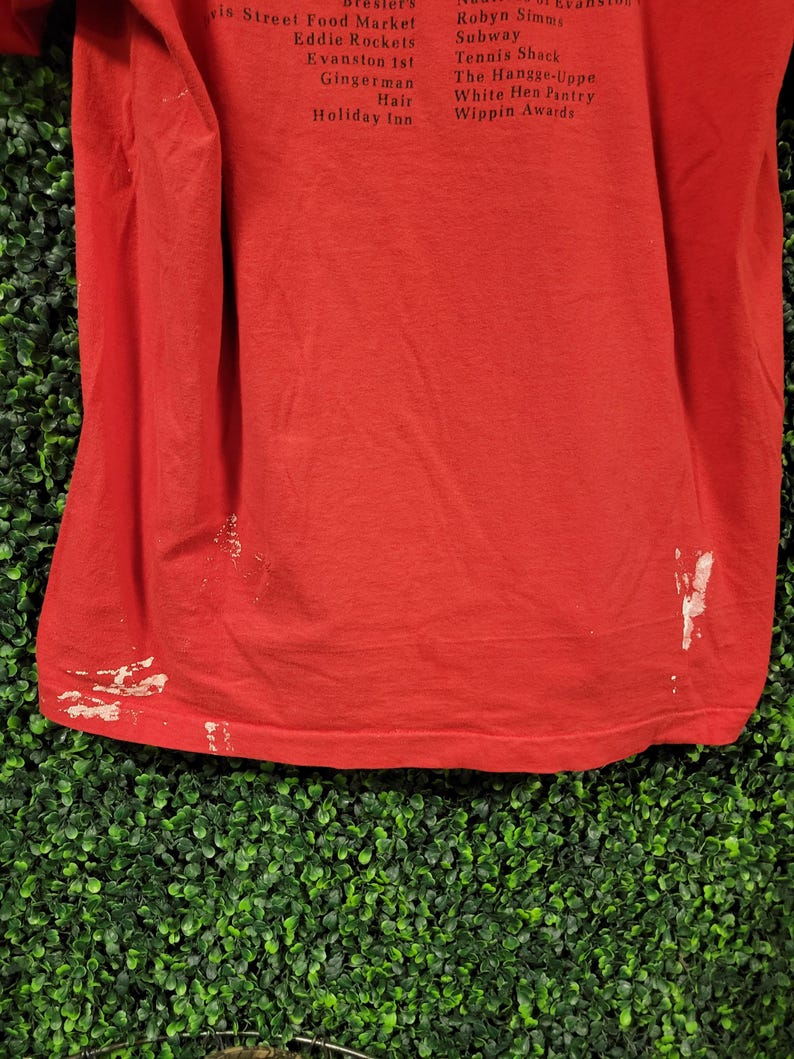 May include: A red t-shirt with white paint marks near the bottom hem. The shirt has a list of text printed on the upper portion. The text includes names of businesses and locations.