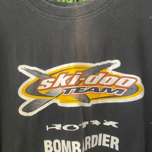 Op de afbeelding: Zwart T-shirt met ronde hals met een afbeelding met de tekst "Ski-Doo TEAM" in witte en rode letters, met een gele en zilveren omtrek. Daaronder staan de woorden "Rotax" en "Bombardier" in het wit.