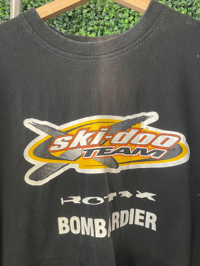 Op de afbeelding: Zwart T-shirt met ronde hals met een afbeelding met de tekst "Ski-Doo TEAM" in witte en rode letters, met een gele en zilveren omtrek. Daaronder staan de woorden "Rotax" en "Bombardier" in het wit.