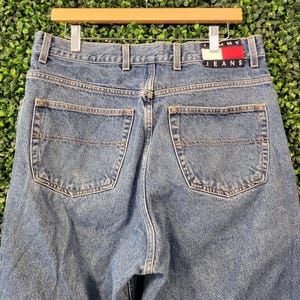 May include: Light blue denim jeans with a classic five-pocket design. The jeans feature a straight leg cut and a red and white Tommy Jeans logo patch on the back pocket. The jeans are hanging on a wooden hanger.