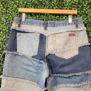 May include: Patchwork denim jeans with a variety of blue and light blue denim panels. The jeans have a frayed hem and pocket details. The back of the jeans is visible, hanging on a wooden hanger.