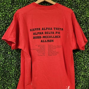 May include: A red t-shirt with black text. The text includes "KAPPA ALPHA THETA", "ALPHA DELTA PHI", "BOBB-MCCULLOCH", and "ALLISON". Additional text lists various names and places. The shirt is hanging on a wooden hanger.