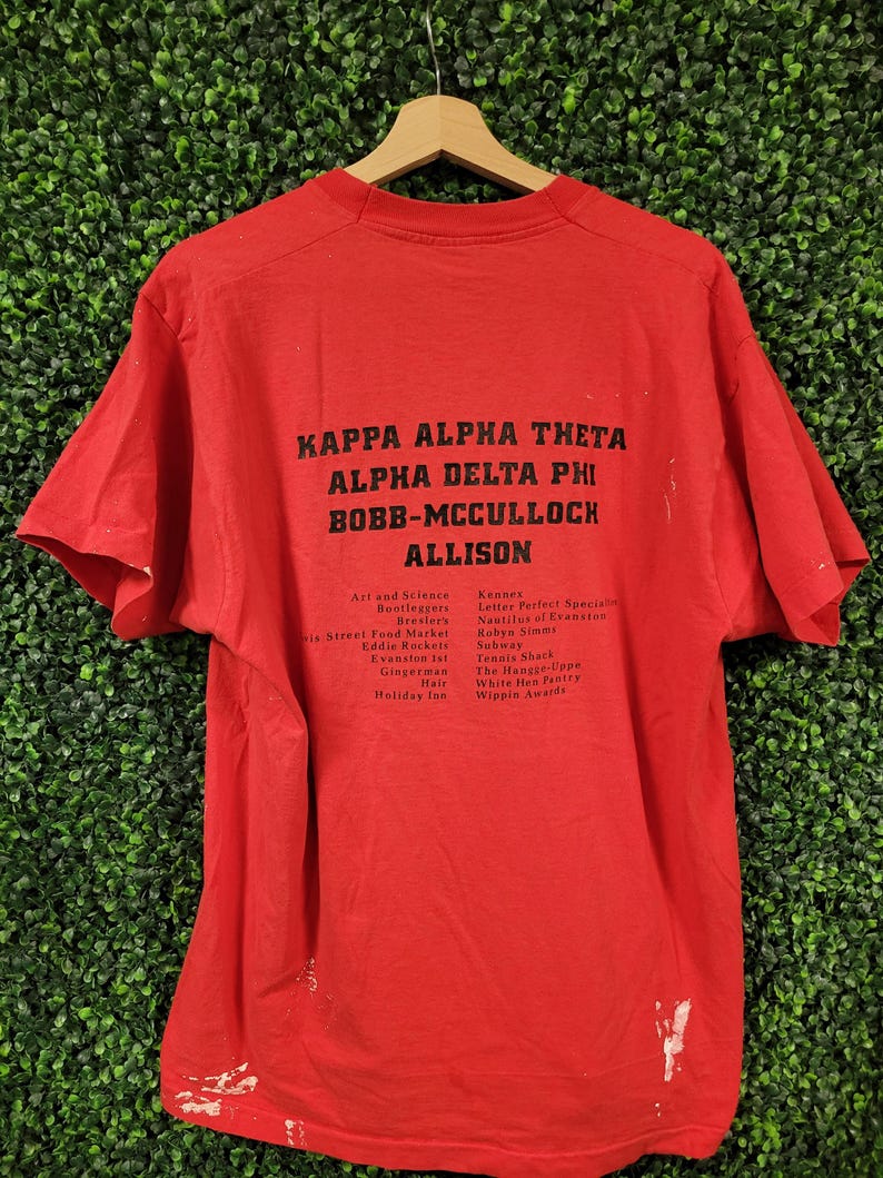 May include: A red t-shirt with black text. The text includes "KAPPA ALPHA THETA", "ALPHA DELTA PHI", "BOBB-MCCULLOCH", and "ALLISON". Additional text lists various names and places. The shirt is hanging on a wooden hanger.