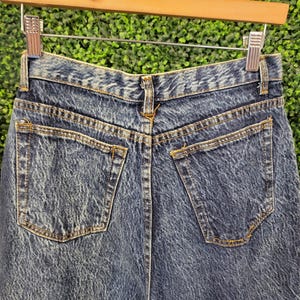 May include: Close-up of the back of acid wash denim jeans. The jeans have two back pockets and a waistband with belt loops. The denim has a faded, textured appearance. The jeans are hanging on a wooden hanger.