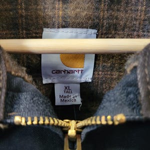 May include: A dark-colored jacket with a brown and black plaid interior lining. The garment features a gold-toned metal zipper and a white interior label with the Carhartt logo, size XL Tall, and the text Made in Mexico printed in black.