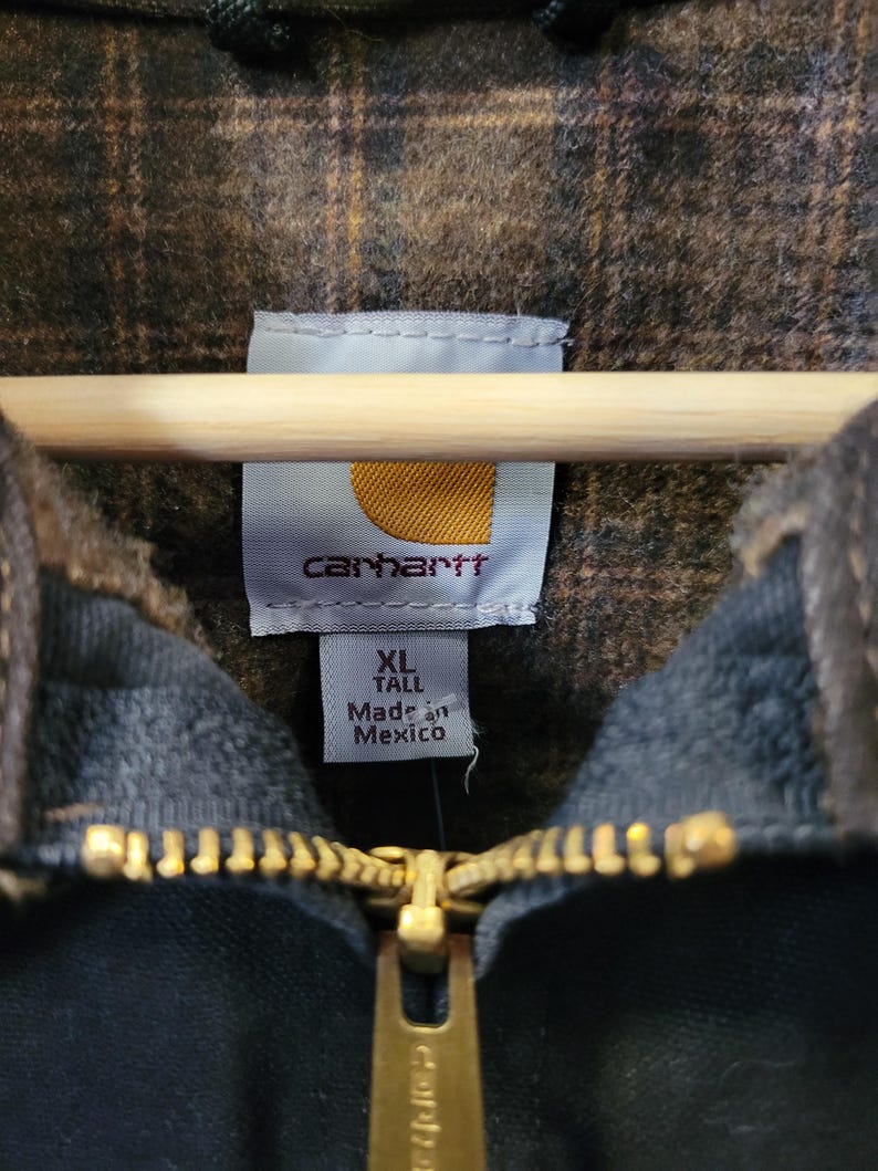 May include: A dark-colored jacket with a brown and black plaid interior lining. The garment features a gold-toned metal zipper and a white interior label with the Carhartt logo, size XL Tall, and the text Made in Mexico printed in black.