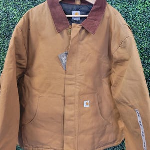 Puede incluir: Una chaqueta Carhartt de color tostado con cuello de pana marrón, forro interior acolchado negro y cierre de cremallera frontal. La chaqueta incluye dos grandes bolsillos delanteros, un parche con el logo de Carhartt y una etiqueta de papel.