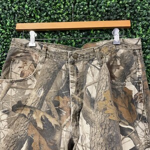 May include: Camouflage-print denim trousers with a button closure, belt loops, and front and back pockets. The trousers feature a realistic print of tree bark and leaves in shades of brown, tan, and green. The trousers are hanging on a wooden hanger.