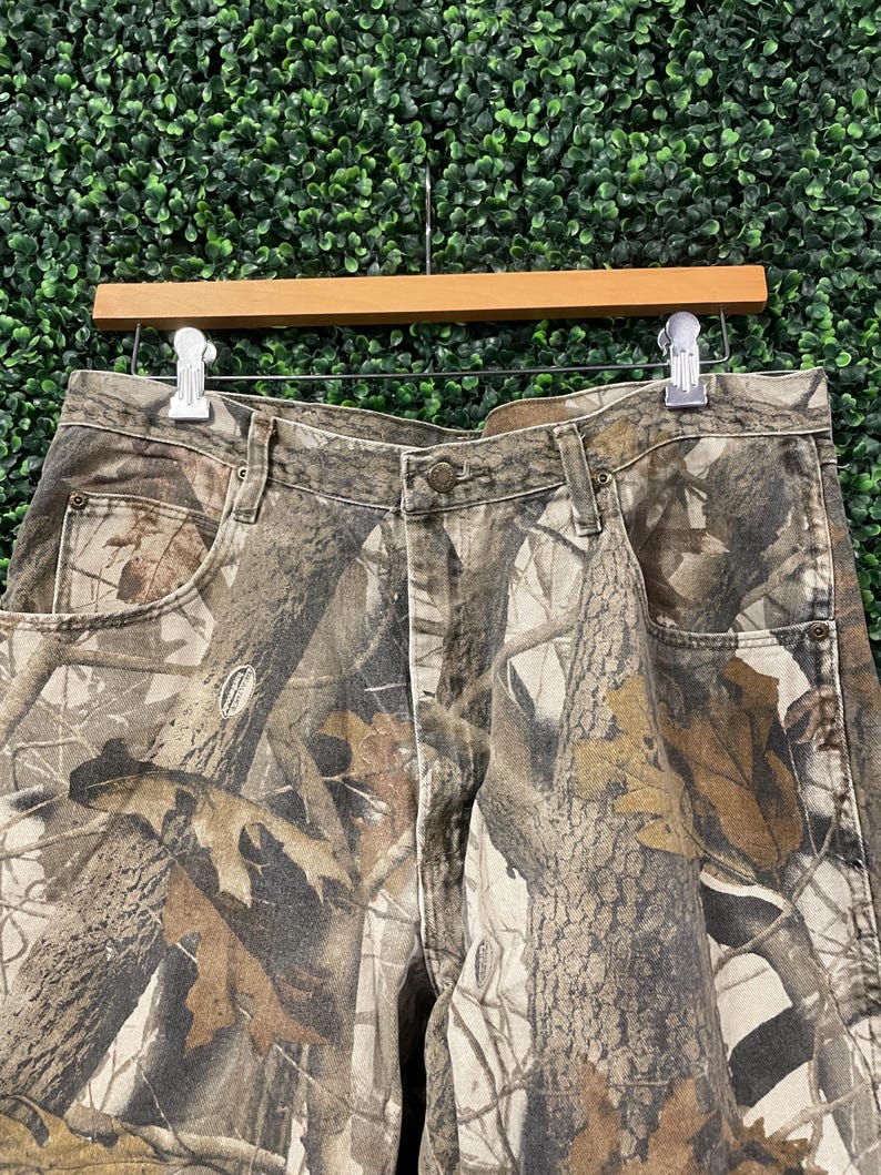 May include: Camouflage-print denim trousers with a button closure, belt loops, and front and back pockets. The trousers feature a realistic print of tree bark and leaves in shades of brown, tan, and green. The trousers are hanging on a wooden hanger.