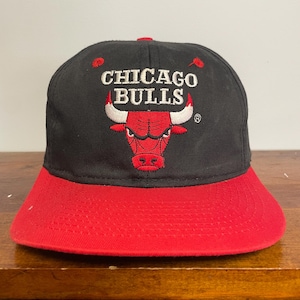 Könnte beinhalten: Eine schwarze Baseballkappe mit rotem Schirm und einem gestickten Chicago Bulls-Logo auf der Vorderseite. Das Logo zeigt einen roten Stierkopf mit weißen Hörnern und den Schriftzug Chicago Bulls in weißen Buchstaben.