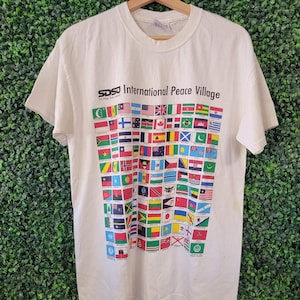 Peut inclure: T-shirt blanc cassé orné d'un motif de drapeaux internationaux colorés. Le texte "SDSU International Peace Village" est imprimé en noir. Le t-shirt est suspendu à un cintre en bois devant un fond vert.
