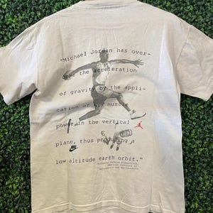 Pu&ograve; includere: T-shirt bianco con grafica in bianco e nero di Michael Jordan in azione, con testo descrittivo. Il logo Nike &egrave; visibile. La maglietta &egrave; appesa a una gruccia di legno.