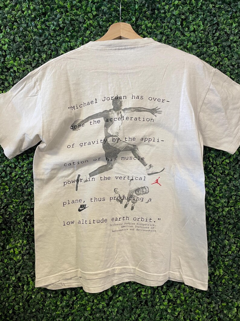 Pu&ograve; includere: T-shirt bianco con grafica in bianco e nero di Michael Jordan in azione, con testo descrittivo. Il logo Nike &egrave; visibile. La maglietta &egrave; appesa a una gruccia di legno.