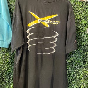 Op de afbeelding: Zwart T-shirt met een geel en wit grafisch ontwerp. Het ontwerp toont het woord "TEAM" met een gele "X" en witte gebogen lijnen. Het T-shirt hangt aan een houten hanger.