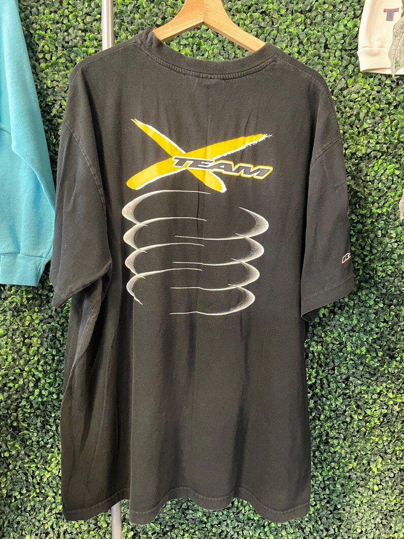 Op de afbeelding: Zwart T-shirt met een geel en wit grafisch ontwerp. Het ontwerp toont het woord "TEAM" met een gele "X" en witte gebogen lijnen. Het T-shirt hangt aan een houten hanger.