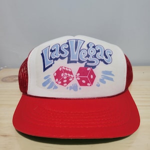 May include: A red and white trucker cap featuring the text Las Vegas in blue lettering above two pink dice. The cap has a white front panel, a red brim, and red mesh side and back panels. The design includes light blue abstract shapes.