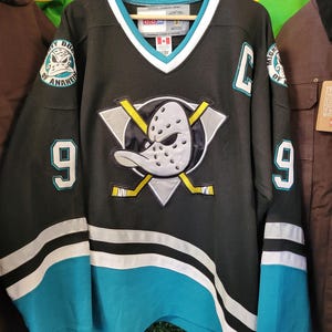 Puede incluir: Jersey de hockey negro con detalles turquesa y blancos. Presenta el logotipo de los Mighty Ducks of Anaheim en el pecho y las mangas, junto con el número 9. El jersey tiene cuello en V y está colgado de una percha de madera.